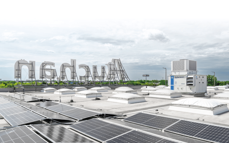 The roof of an Auchan store featuring solar panels, skylights, and a white Rooftop Retrofit IntrCooll unit. The large Auchan logo is visible in the background.