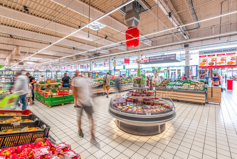 Overzicht van de groente- en fruitafdeling in een Auchan-supermarkt met winkelende klanten, een ronde koeltafel en rode luchtverdeelelementen van een IntrCooll-unit aan het plafond.