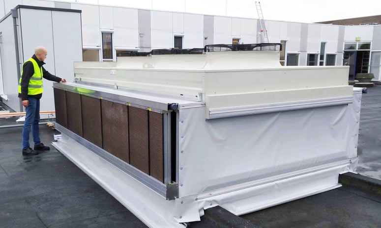 Monteur in veiligheidsvestje inspecteert een grote PreCooll-voorkoelingsunit op een plat dak, met filtermatten aan de zijkant en ventilatoren bovenop.
